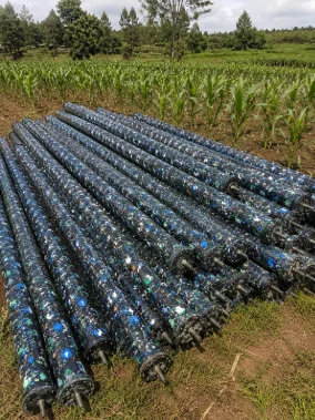 A stack of long, speckled poles made from recycled plastic lies on the ground beside a green field of young corn.
