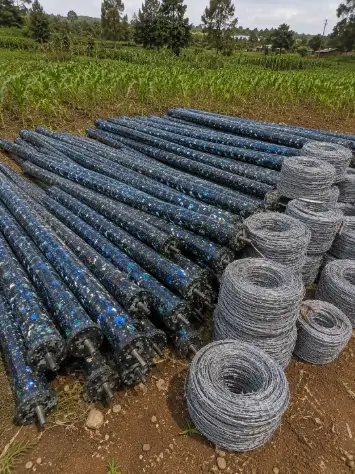 A stack of long, blue-flecked recycled plastic poles lies next to several rolls of barbed wire on a farm, with green crops and trees in the background.