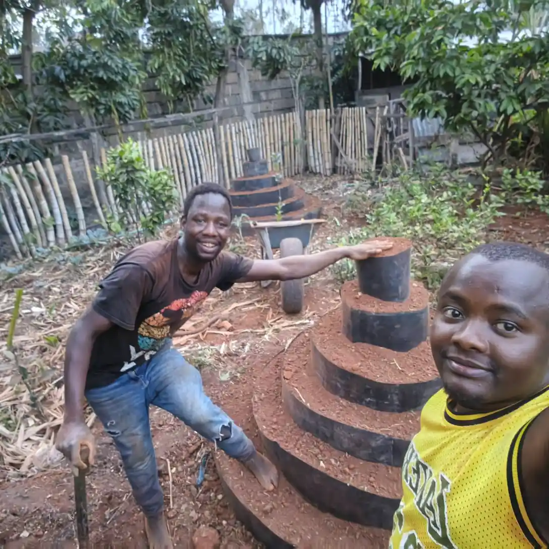 Two men smile for a selfie while building circular, multi-storey vertical garden tower in a lush backyard.